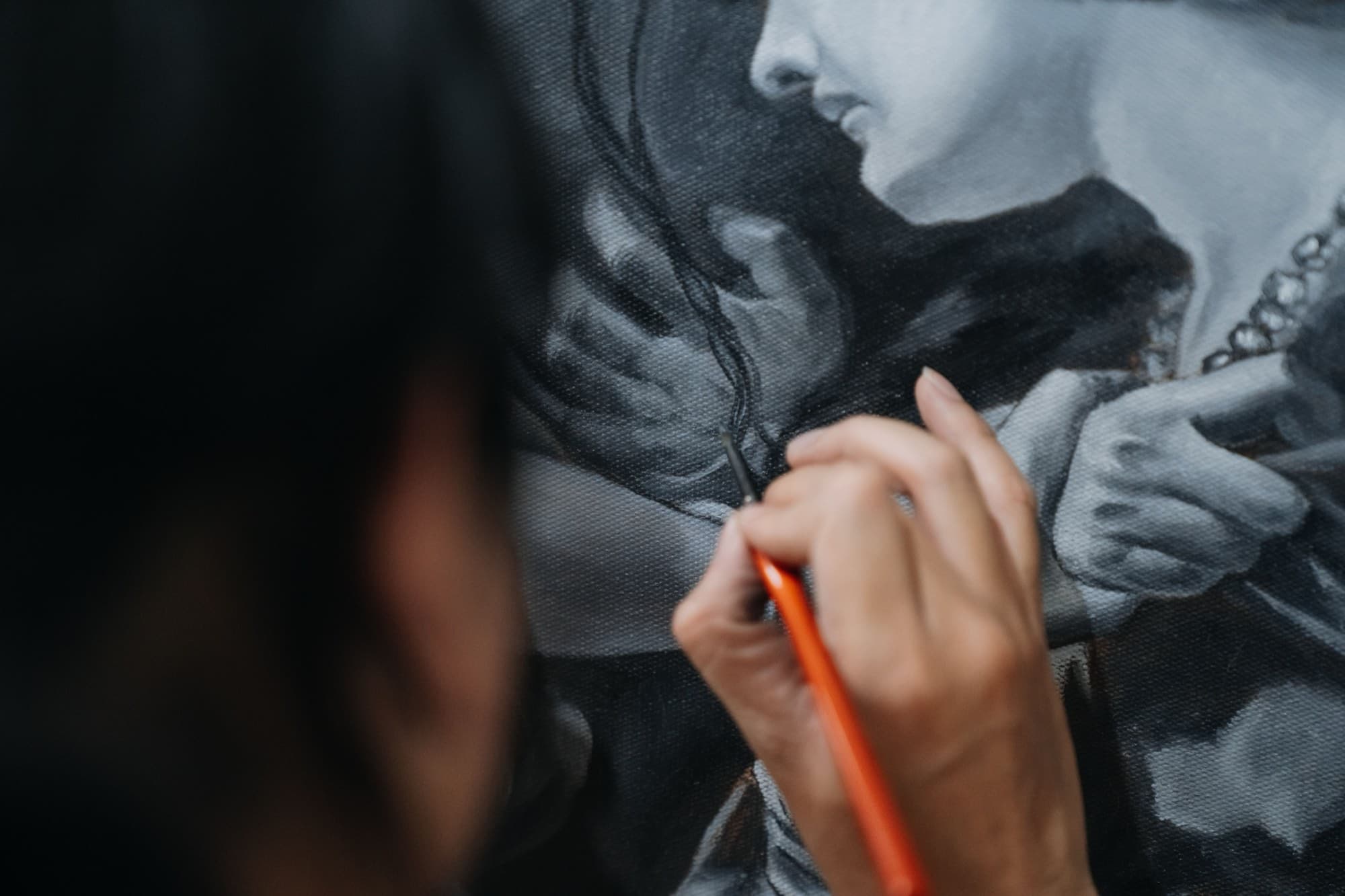 An artist leaning in close to paint fine details on a grayscale portrait canvas with a small brush