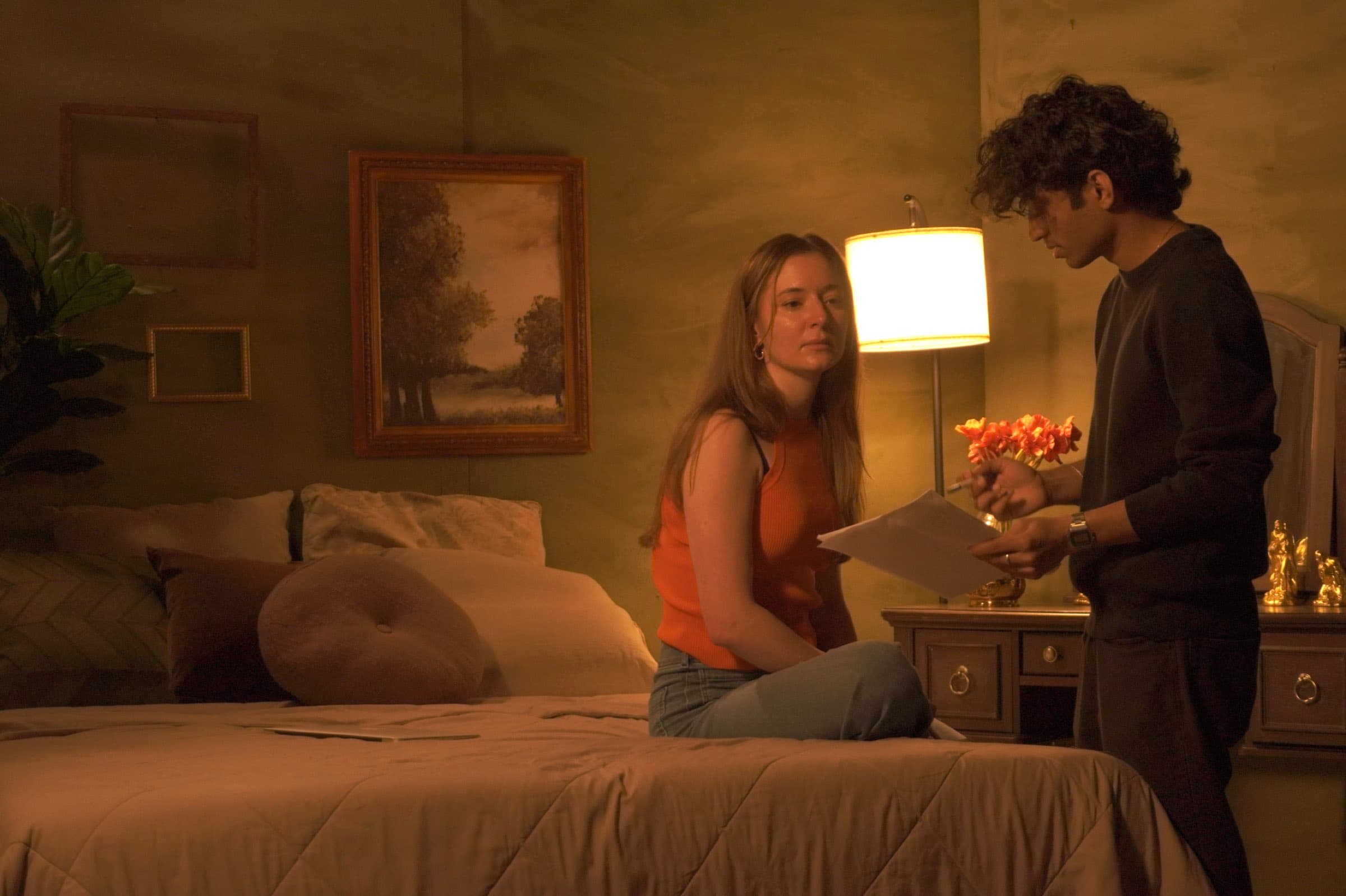A warmly lit bedroom film set with a filmmaker directing two actors, one seated on the bed holding a script and the other standing with a bouquet of flowers