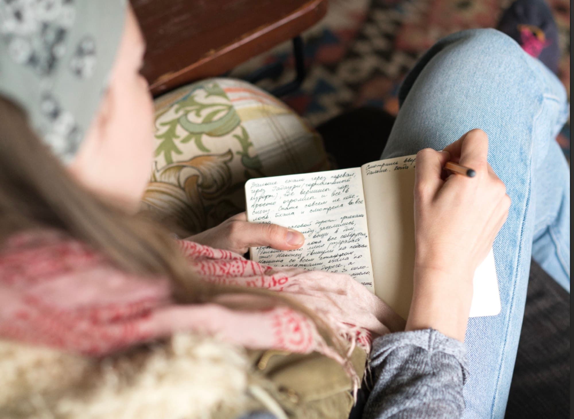 A person sitting cross-legged, handwriting notes in an open journal while holding a pencil