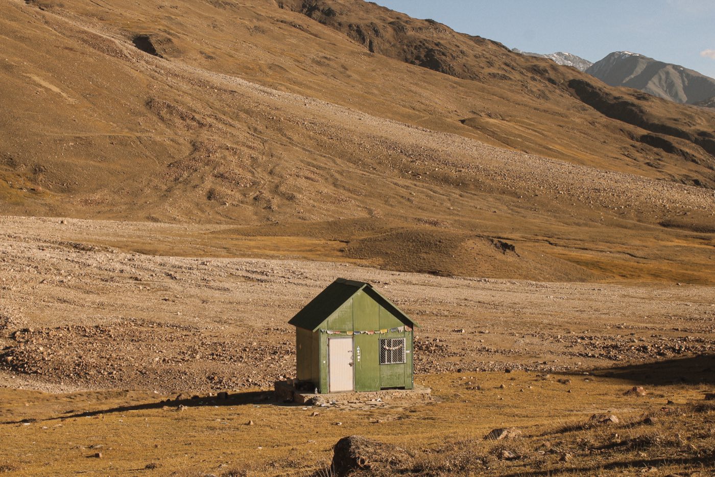 A small green hut standing alone in a cold desert landscape, framed by bare mountains