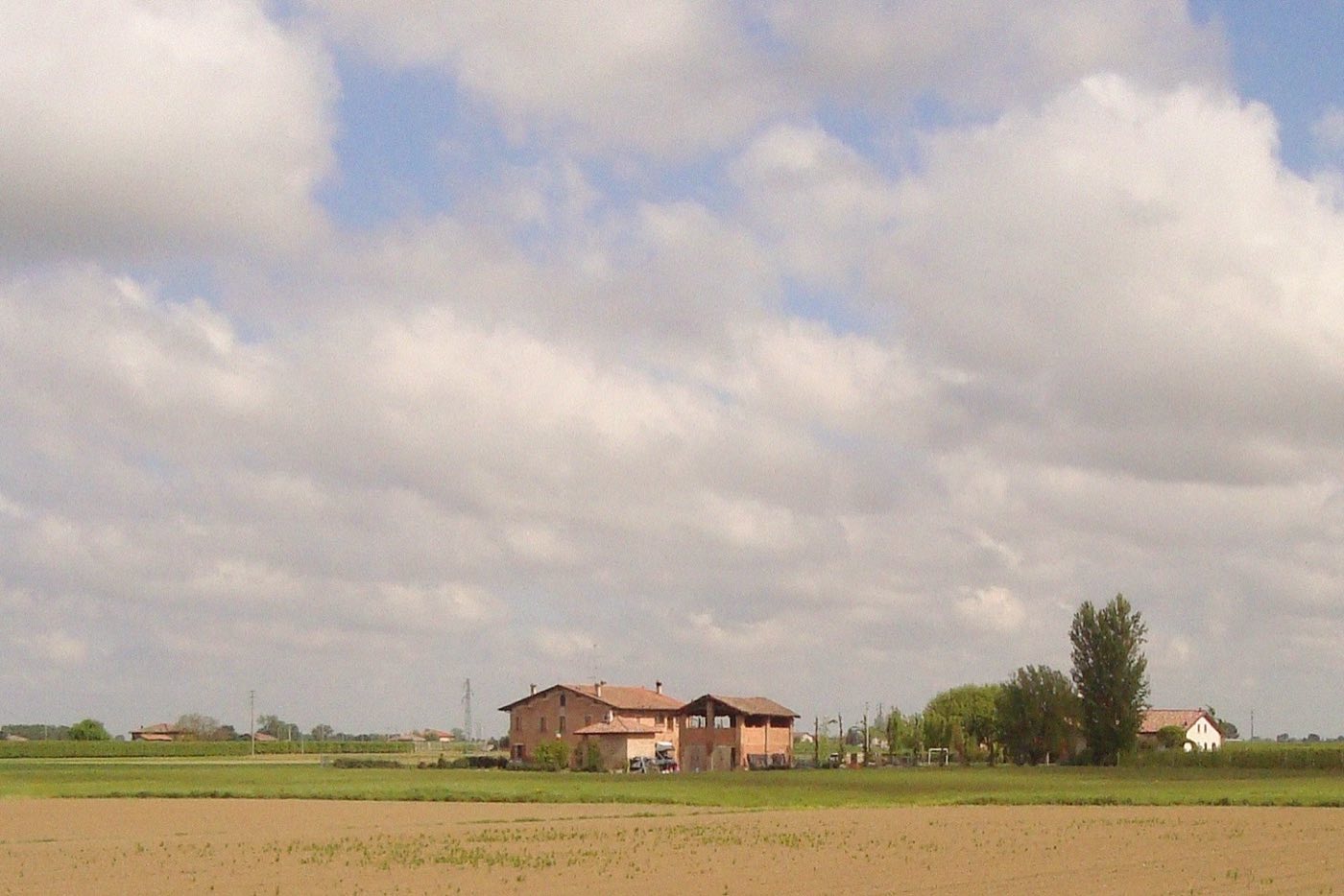 A farmhouse surrounded by green fields under a wide, cloud-filled sky, taken on a trip to Italy
