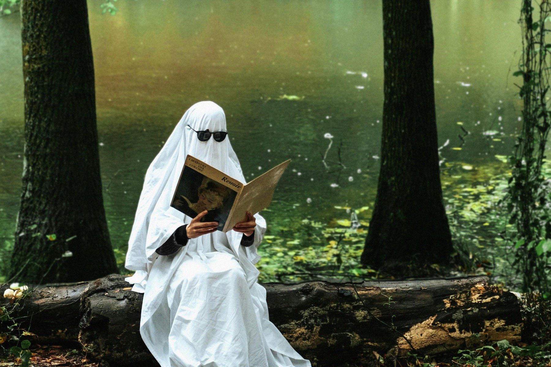 A figure in a white ghost costume and sunglasses sits on a log by a green pond, reading a Renoir art book