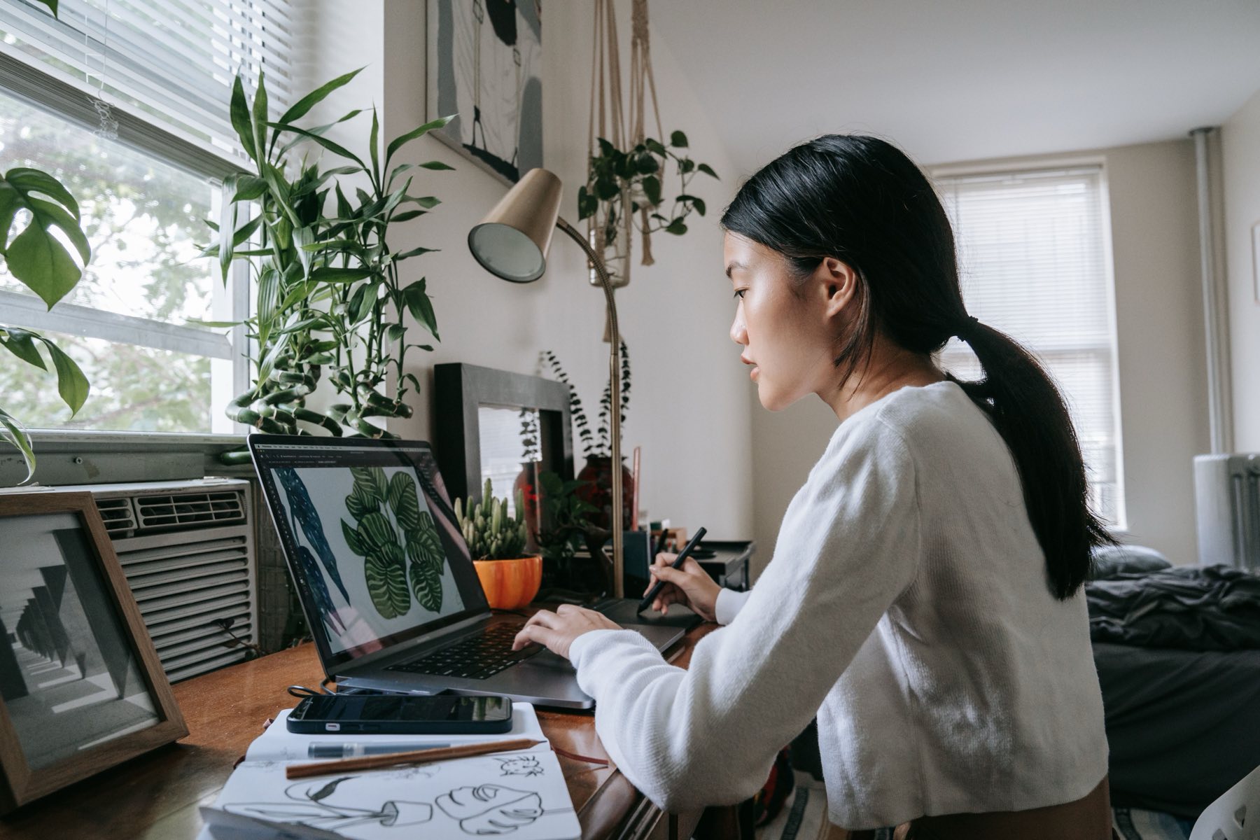 An artist working at a laptop and drawing tablet in a plant-filled home studio, sketches and a sketchbook open on the desk beside her