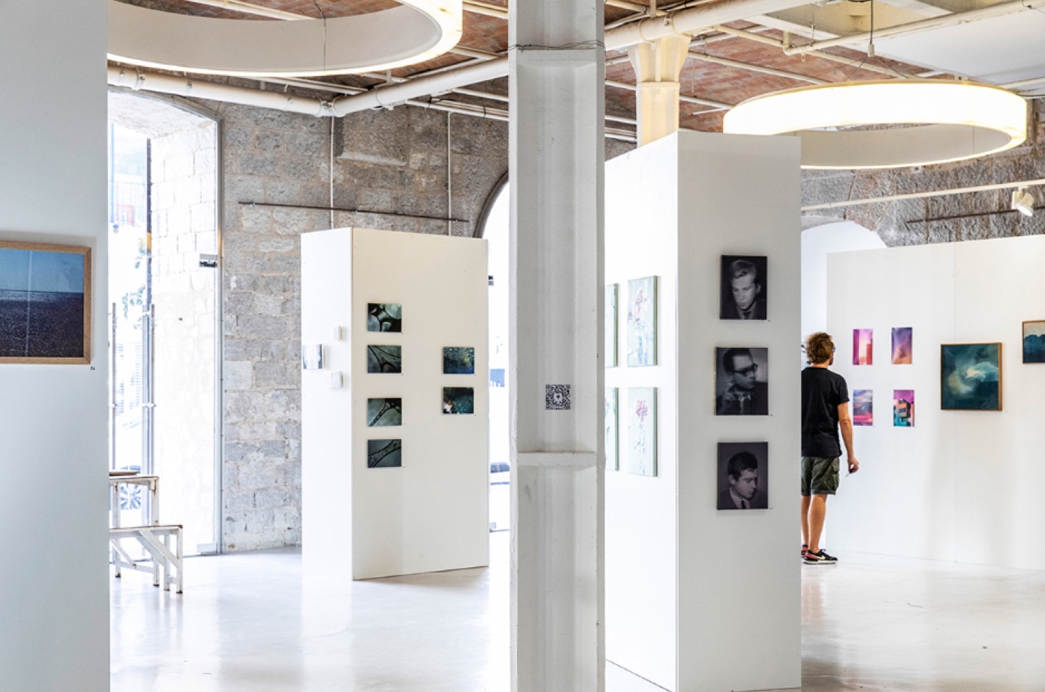 Interior view of a gallery installation space with white partition walls displaying framed photographs, a visitor viewing portraits in the background
