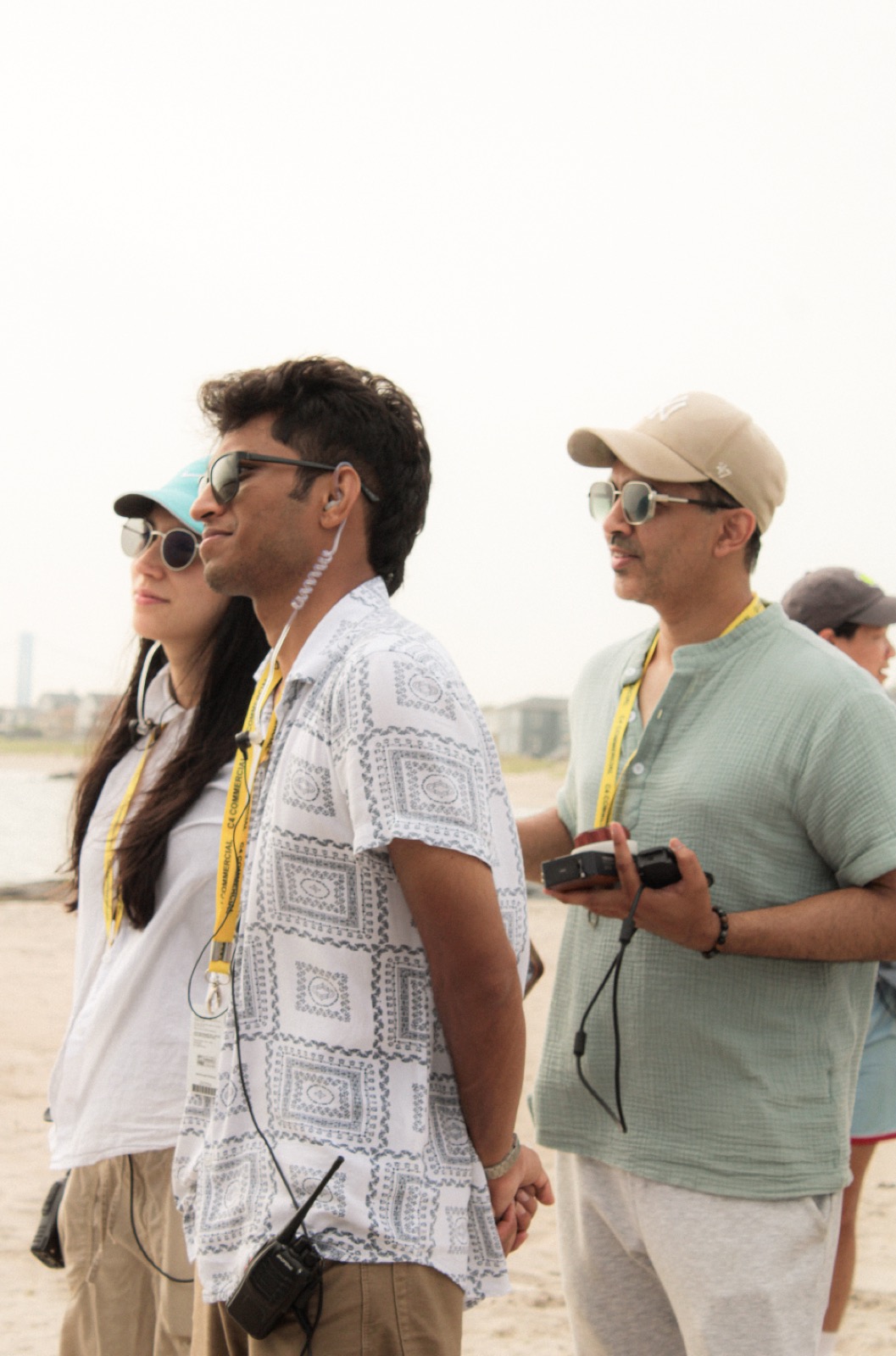 Nived Krishna standing with two crew members on a beach during a commercial shoot, wearing headphones and holding a walkie-talkie