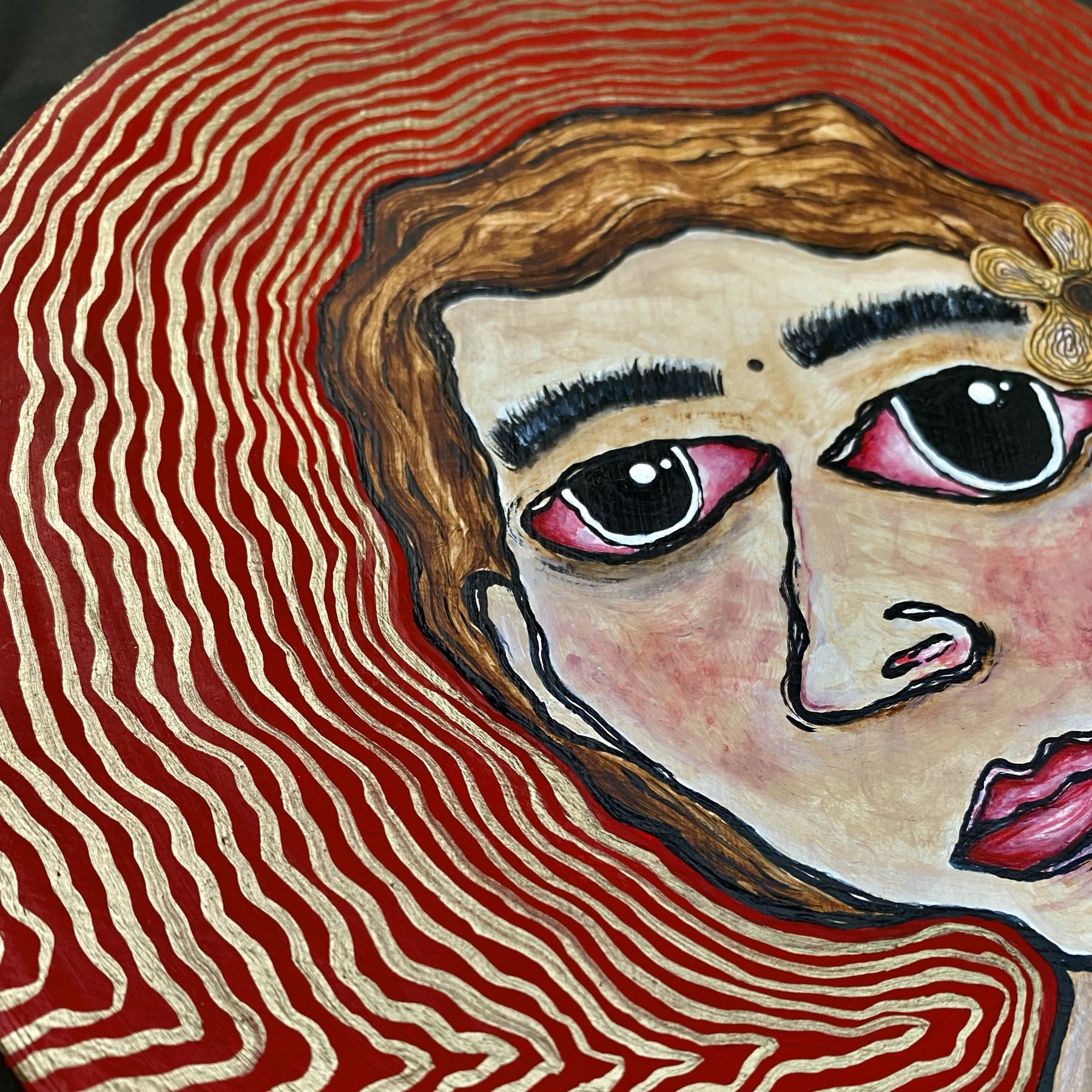Close-up detail of a hand-painted circular wooden artwork showing a woman's face surrounded by flowing red and gold wave patterns